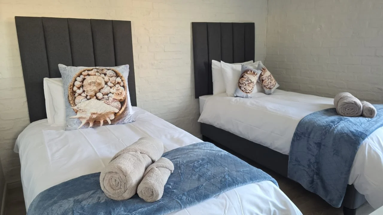 Two beds with black headboards, white bedding, blue bed runners, rolled towels, and decorative pillows with shell designs, against a white brick wall.