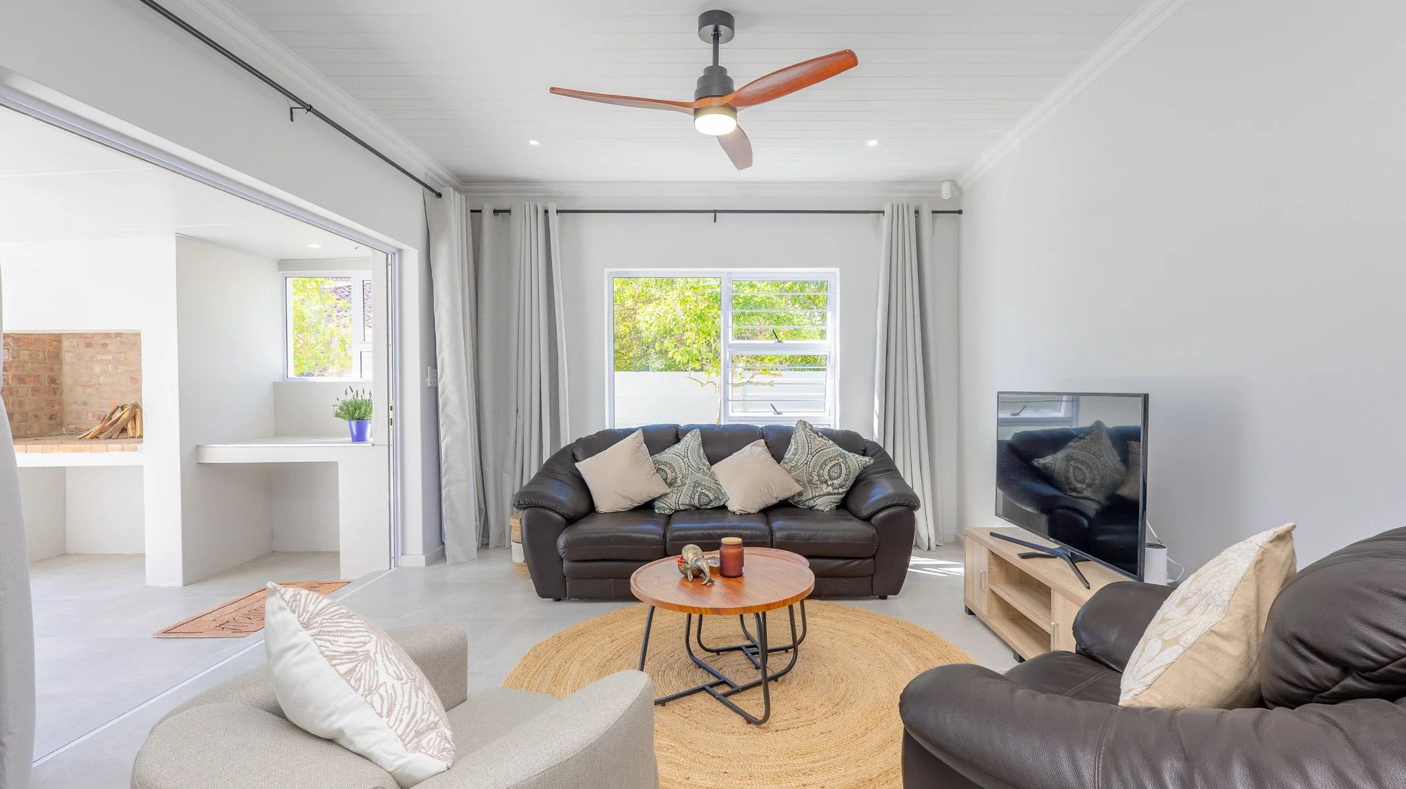 Bright living room with white walls, a ceiling fan, a black leather sofa with decorative pillows, a wooden coffee table, and a flat-screen TV on a light wood stand. There are large windows with white curtains, and a cozy beige armchair with pillows. 