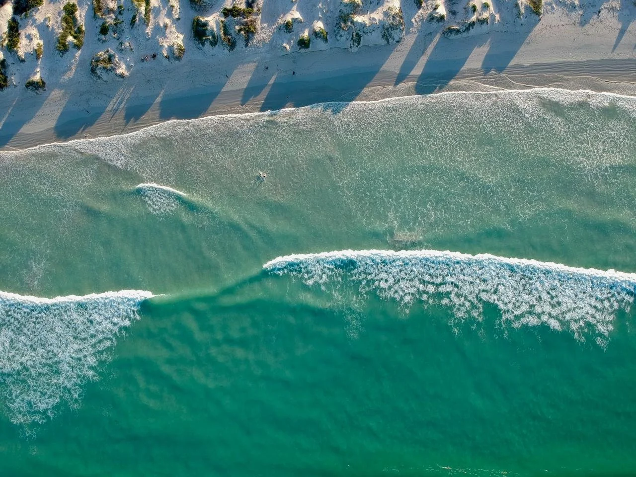 Aerial view of a beach with greenish-blue ocean waves crashing onto the sandy shoreline, with rocky cliffs and trees along the coast casting shadows.