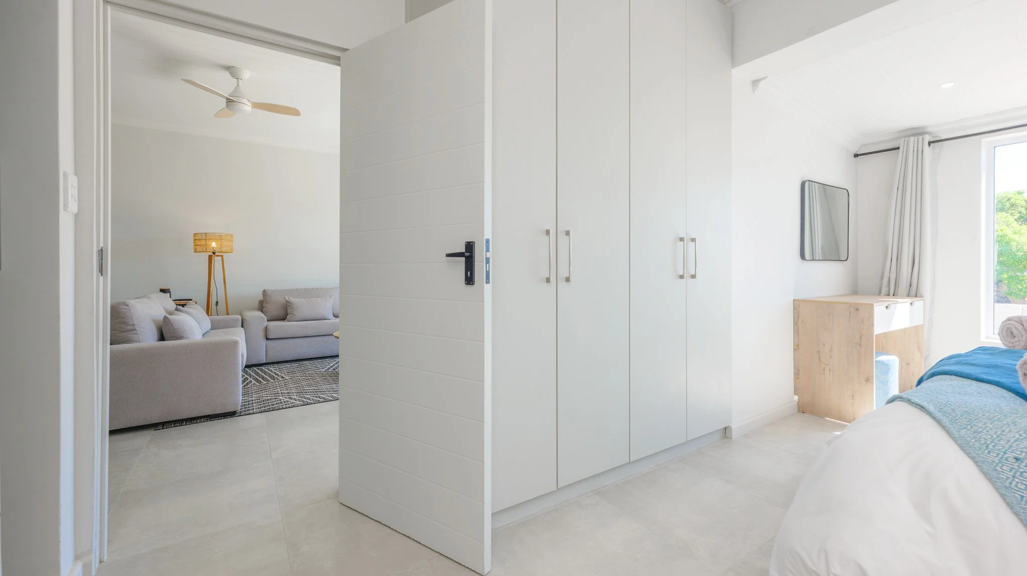 Interior view of a modern bedroom with a white wardrobe, a bed with blue linens, a small wooden vanity or desk, a window with white curtains, and a sitting area with beige sofas and a standing floor lamp with a woven shade.