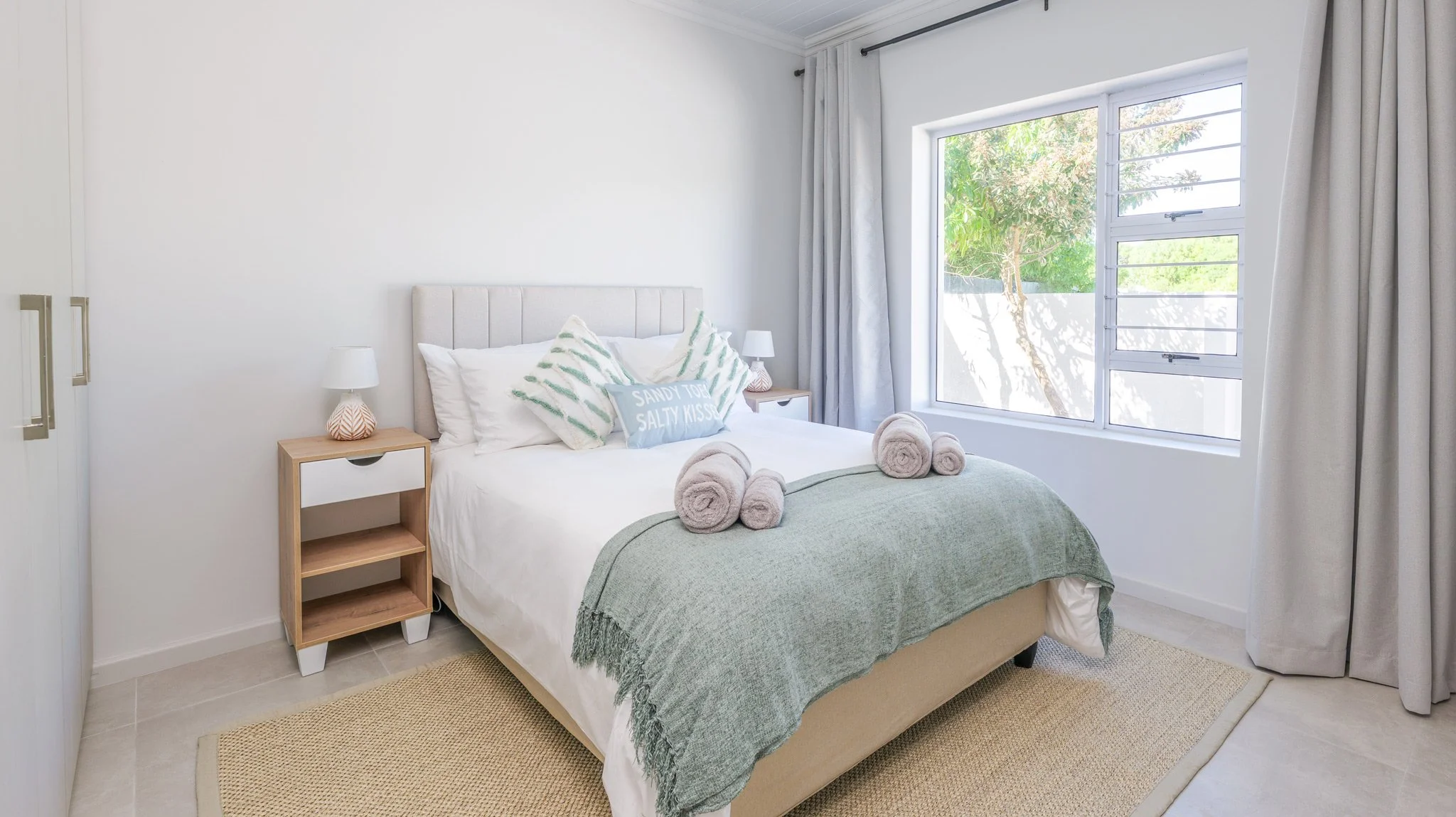 A neatly made bedroom with a white bed, gray pillows, and rolled towels on top. The room has a large window with sheer curtains, and two small bedside lamps on wooden nightstands.