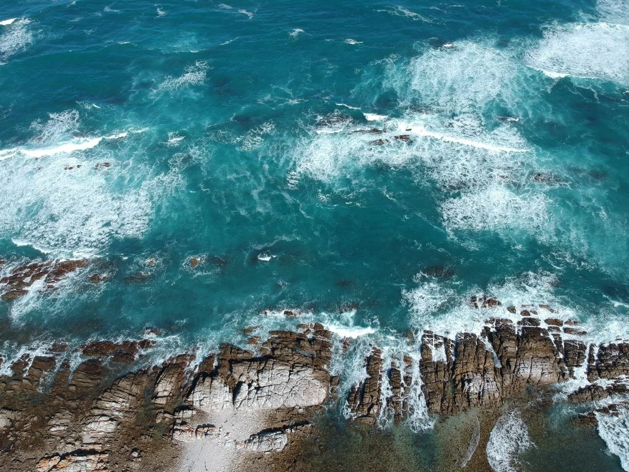 Overhead view of turquoise ocean waves crashing against rocky shoreline.
