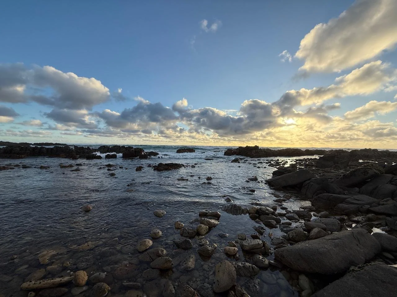 Sunset over a rocky beach with clouds in the sky and gentle waves in the water.