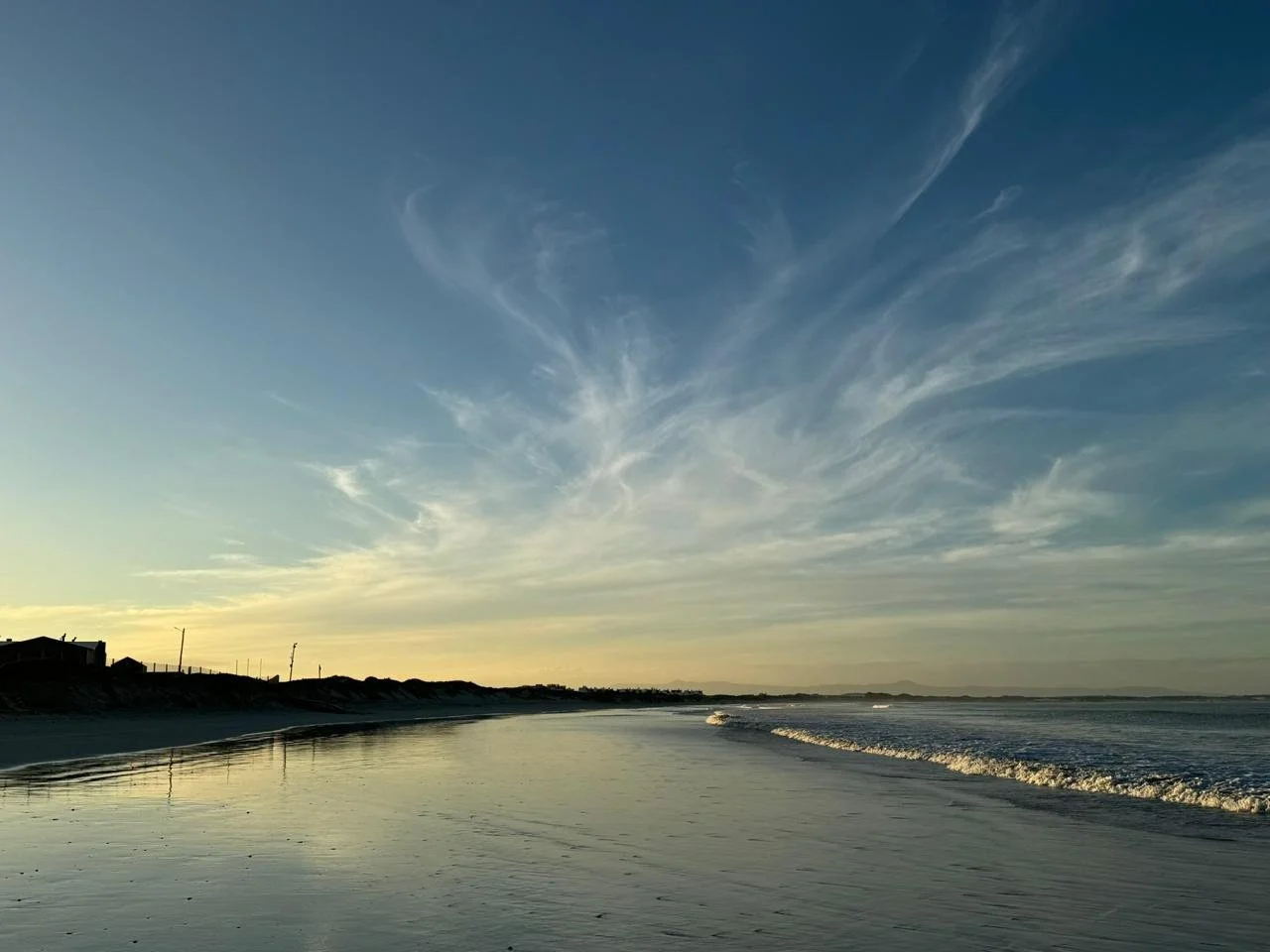 Sunset over a calm beach with a clear sky, wispy clouds, and a shoreline with gentle waves.