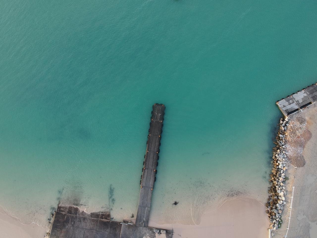 Aerial view of a pier extending into a turquoise ocean, with a sandy beach and rocky shoreline on the right side.