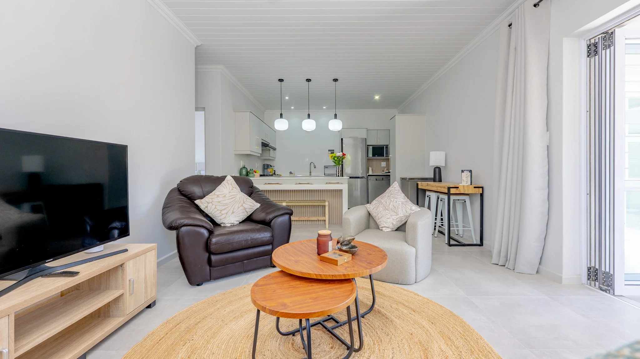 Living room with a black leather armchair and a beige armchair, coffee tables, a TV on a wooden stand, and a view of the kitchen with pendant lights, a refrigerator, and a kitchen island. There are curtains and sliding glass doors to the right.