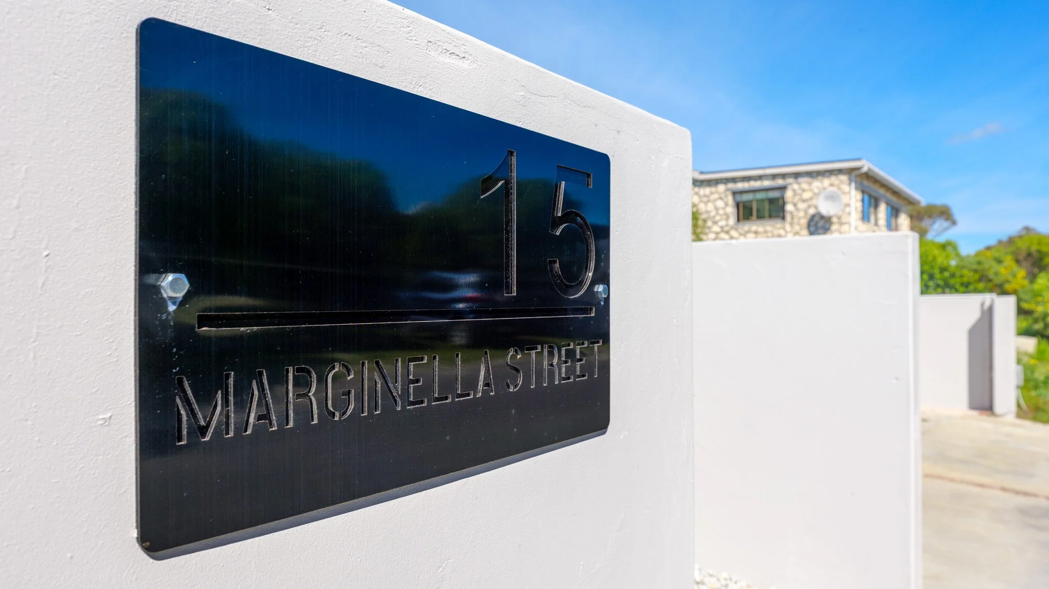 House number 15 on a black reflective mailbox mounted on a white wall, with a house, trees, and a blue sky in the background.