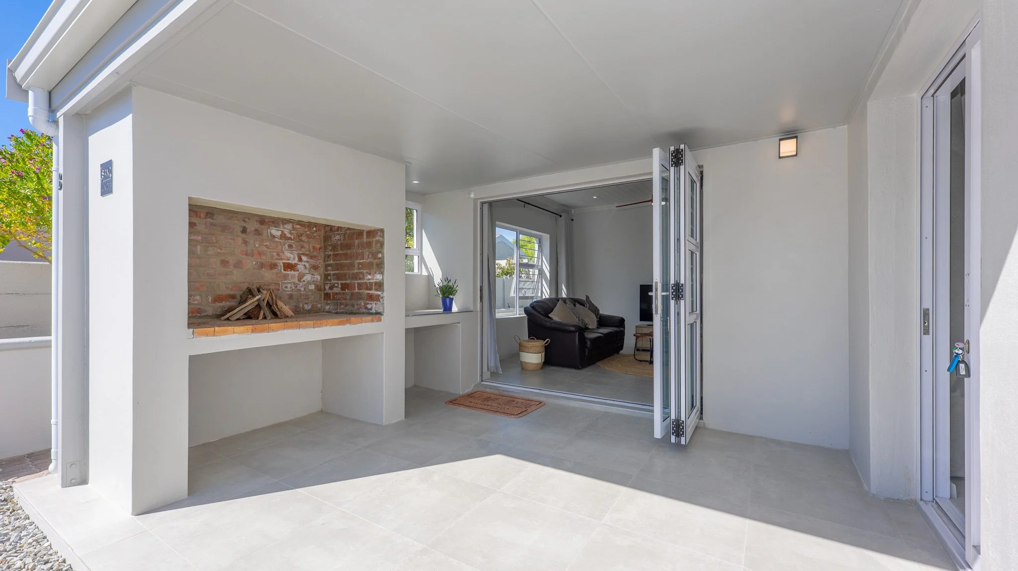 Outdoor patio with built-in brick fireplace, light-colored tiled flooring, white walls, and a view into a living room with a black sofa and windows.
