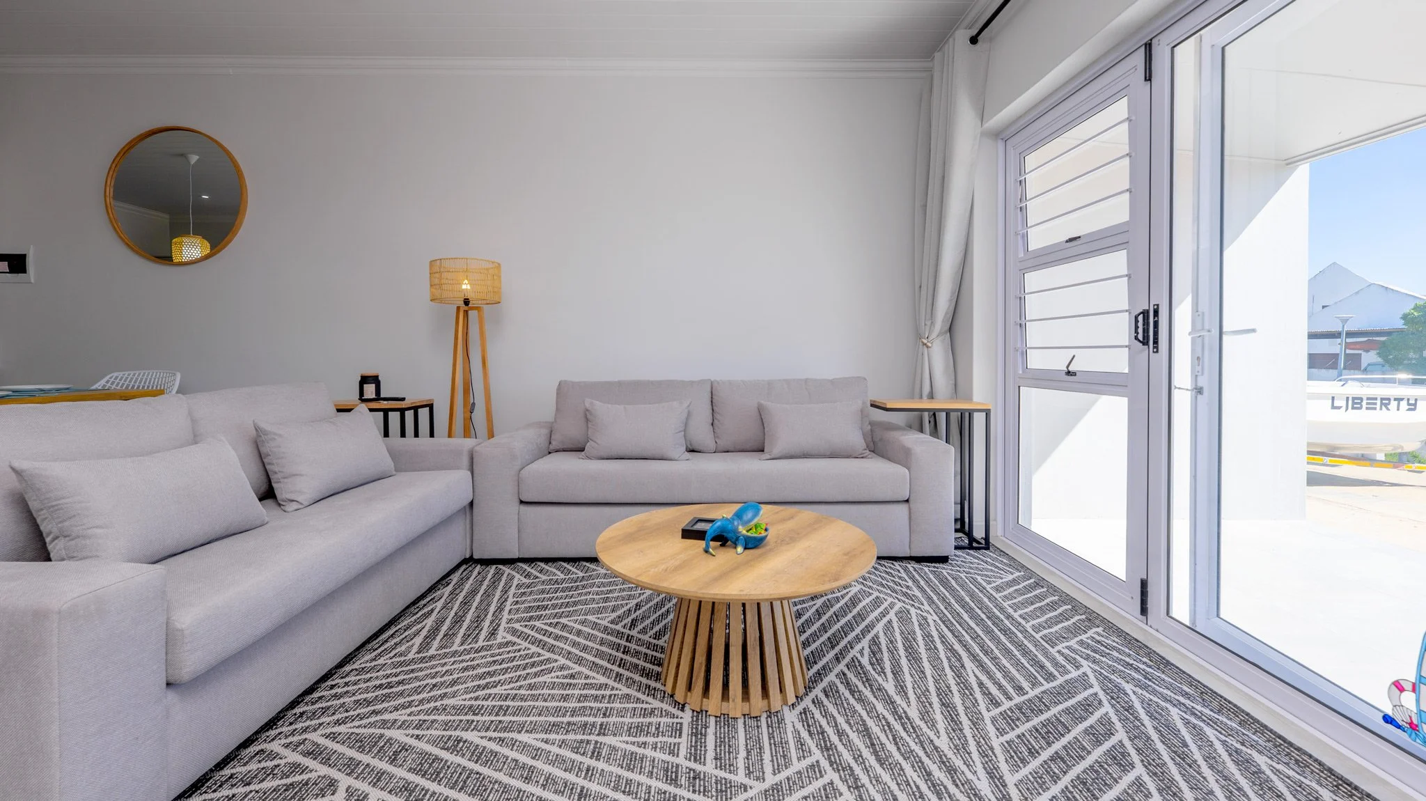 Living room with light-colored sofas, a round wooden coffee table with a blue toy and a black device, patterned rug, floor lamp, mirror on the wall, large sliding glass door with white curtains, and a view of an outdoor boat with the word "LIBERTY".