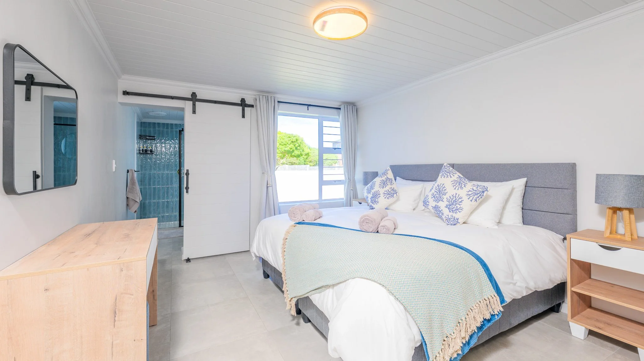 Bright bedroom with a large bed decorated with white and blue patterned pillows, a beige blanket, and three rolled towels. There is a window with white curtains, a wooden nightstand with a gray lamp, a wooden dresser, and a barn-style sliding door th