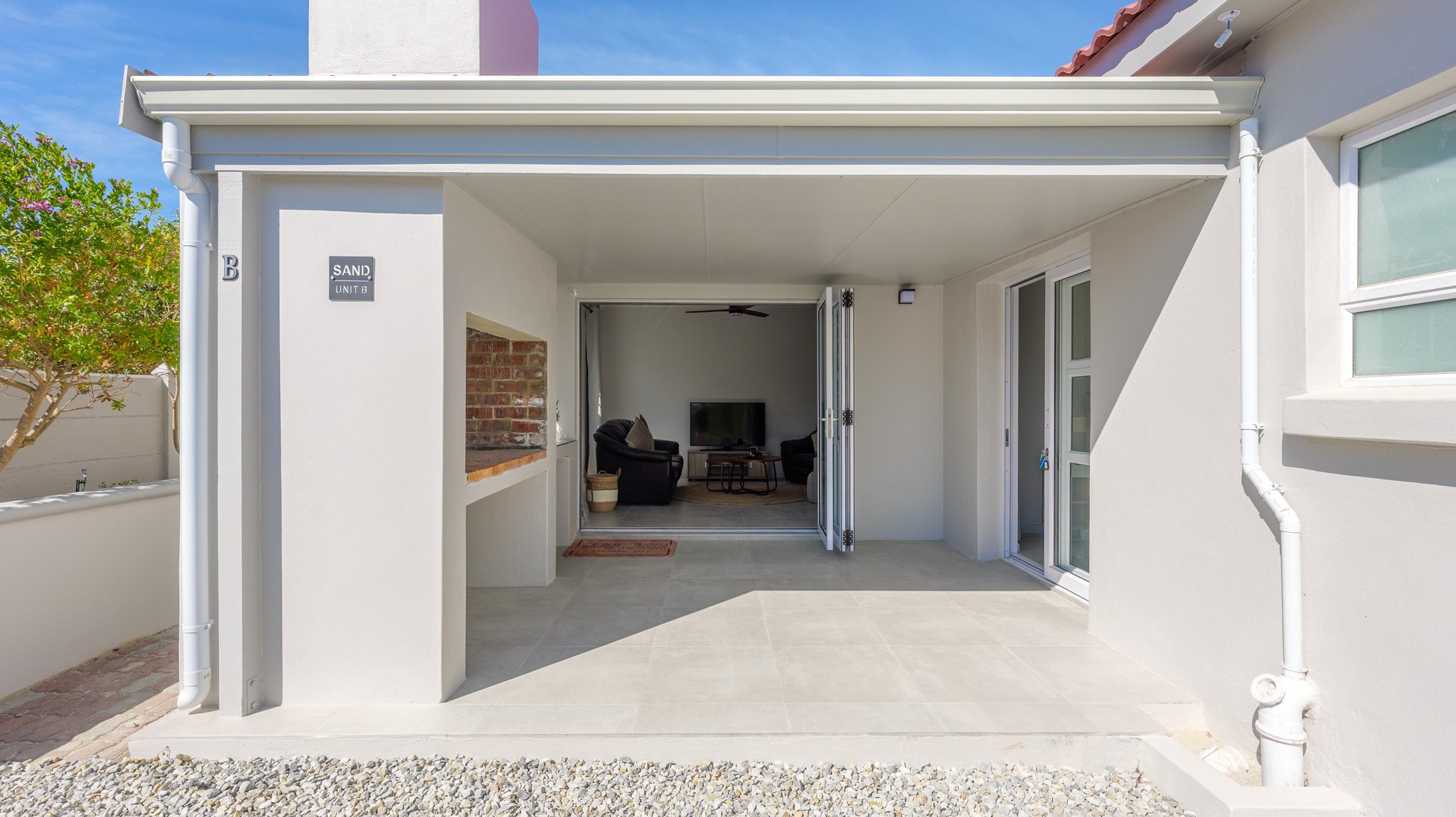 Covered patio area with view into living room with two dark recliners and a flat-screen TV, sliding glass doors, white walls, tiled flooring, and outdoor gravel area.