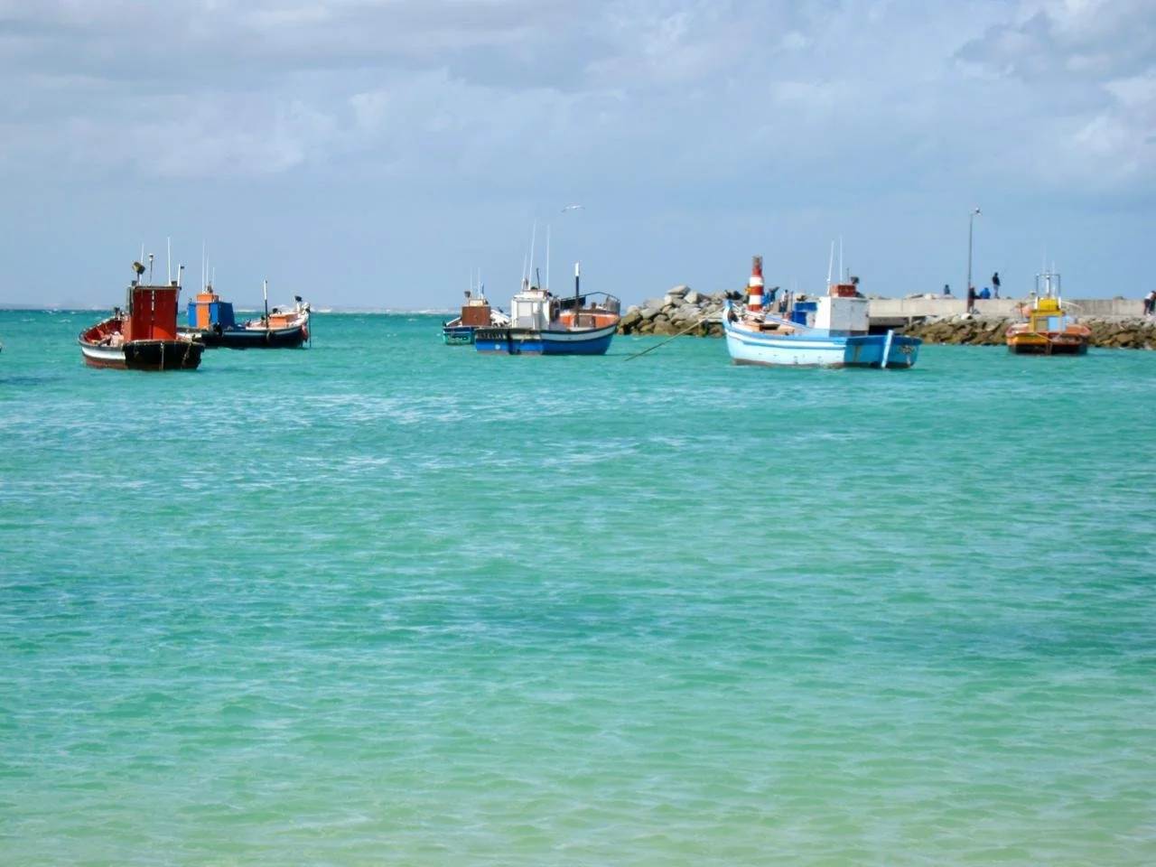 Several small boats anchored near a rocky pier on the turquoise water of the ocean under a partly cloudy sky.