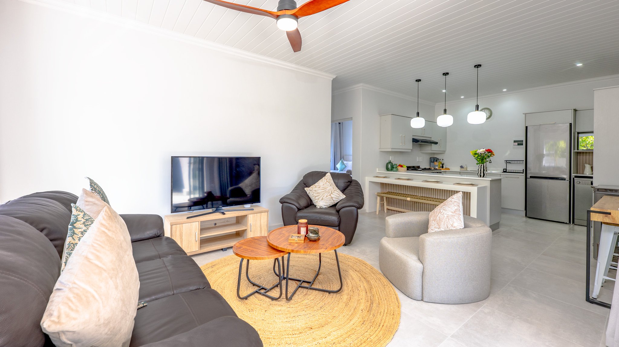 Living room with black leather sofa, beige pillows, small wooden coffee tables, a gray armchair with a beige pillow, TV on wooden stand, white ceiling with ceiling fan, open kitchen with pendant lights, stainless steel refrigerator, white cabinetry, 