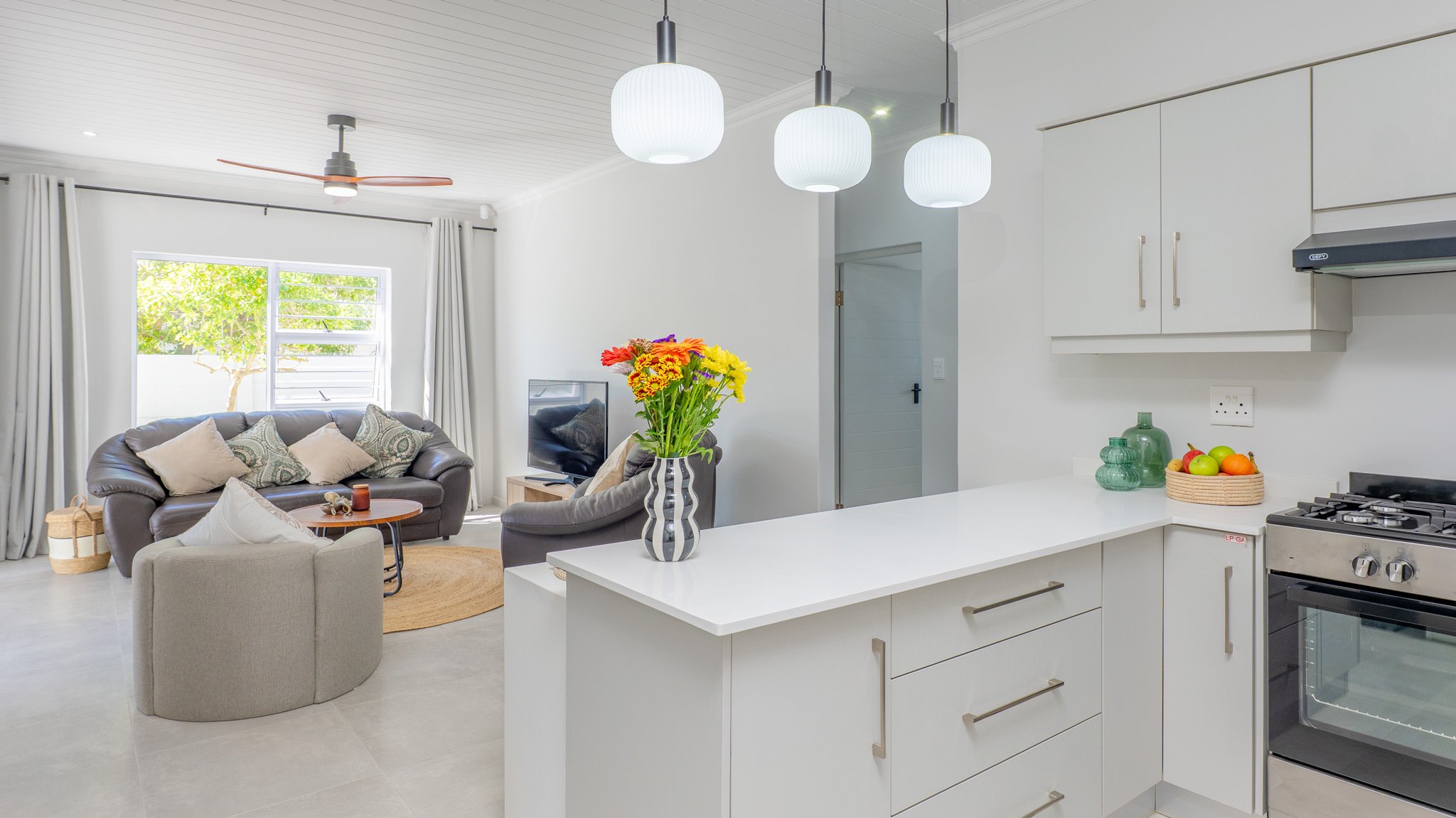 Open-concept living room and kitchen with white cabinets, a vase of colorful flowers, and modern lighting fixtures.