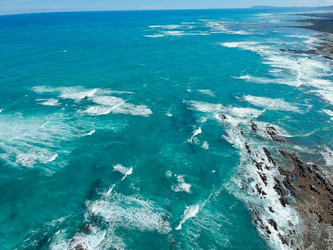 Aerial view of a turquoise ocean with waves crashing against rocky shoreline, stretching to the horizon under a clear sky.