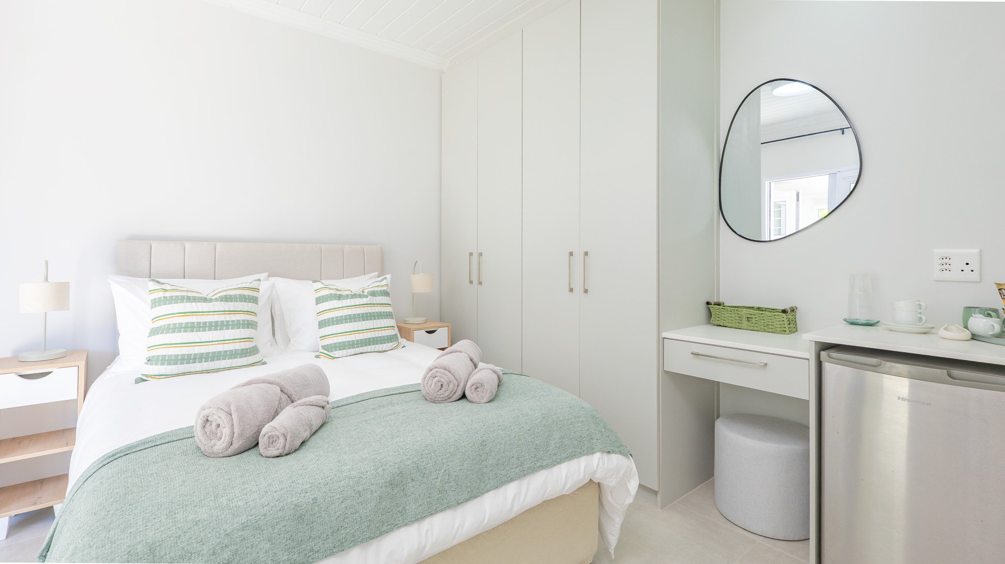 A bedroom with a bed, pillows, and rolled towels, white closet doors, and a small kitchenette with a mini-fridge, a mirror, and decorative items on the counter.