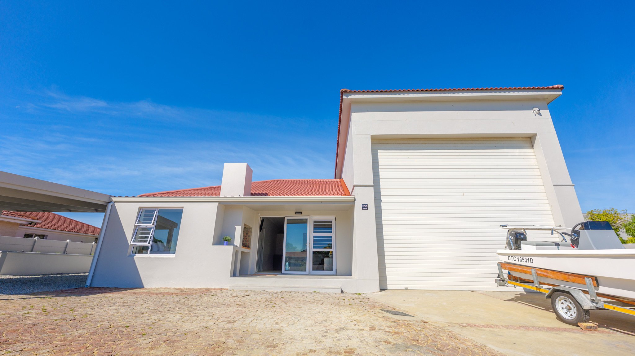Modern white house with a red-tiled roof and a garage door, a small boat on a trailer parked outside, under a clear blue sky.