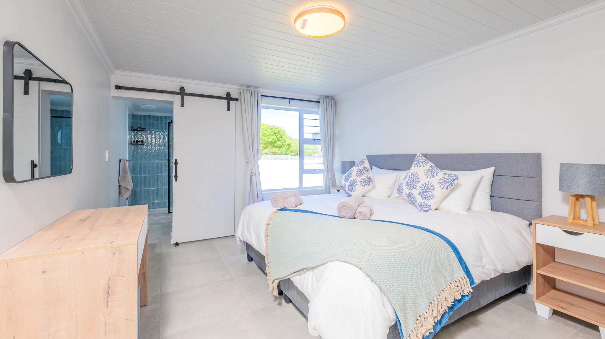 A bright bedroom with a large bed, soft pillows, and a window showing green trees outside, with a view of a bathroom through an open door.