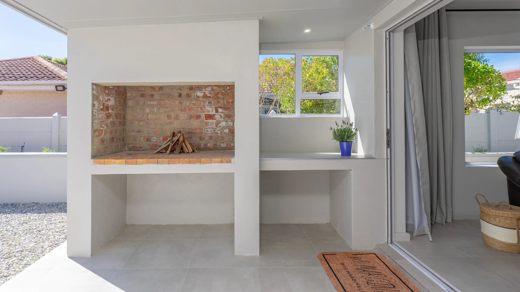 Indoor space with a built-in brick fireplace, a window with a potted plant, and an open sliding door leading outside, with a doormat near the door.