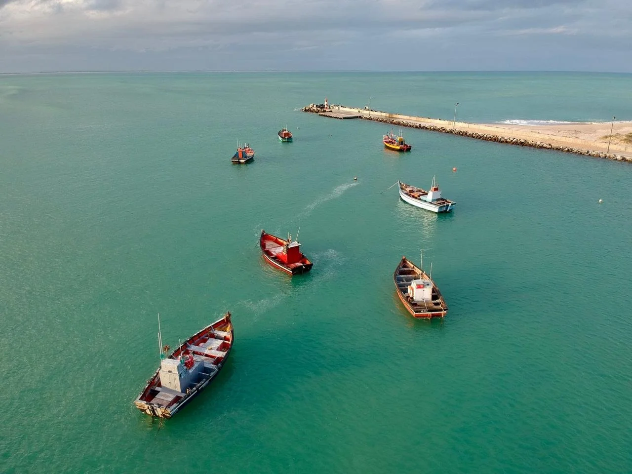 Several boats anchored in a harbor near a breakwater with a lighthouse at the end, under a cloudy sky.