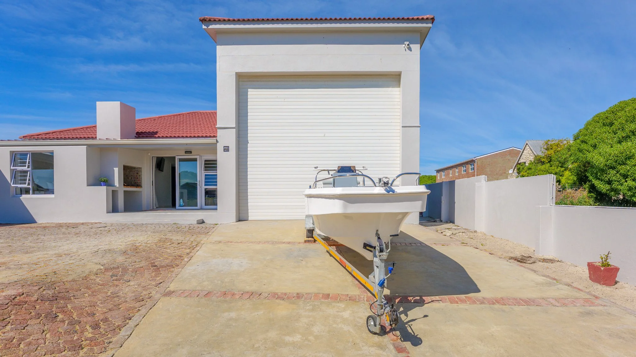 A white building with a large roller shutter door, a boat on a trailer, and a paved driveway with brick accents under a blue sky.