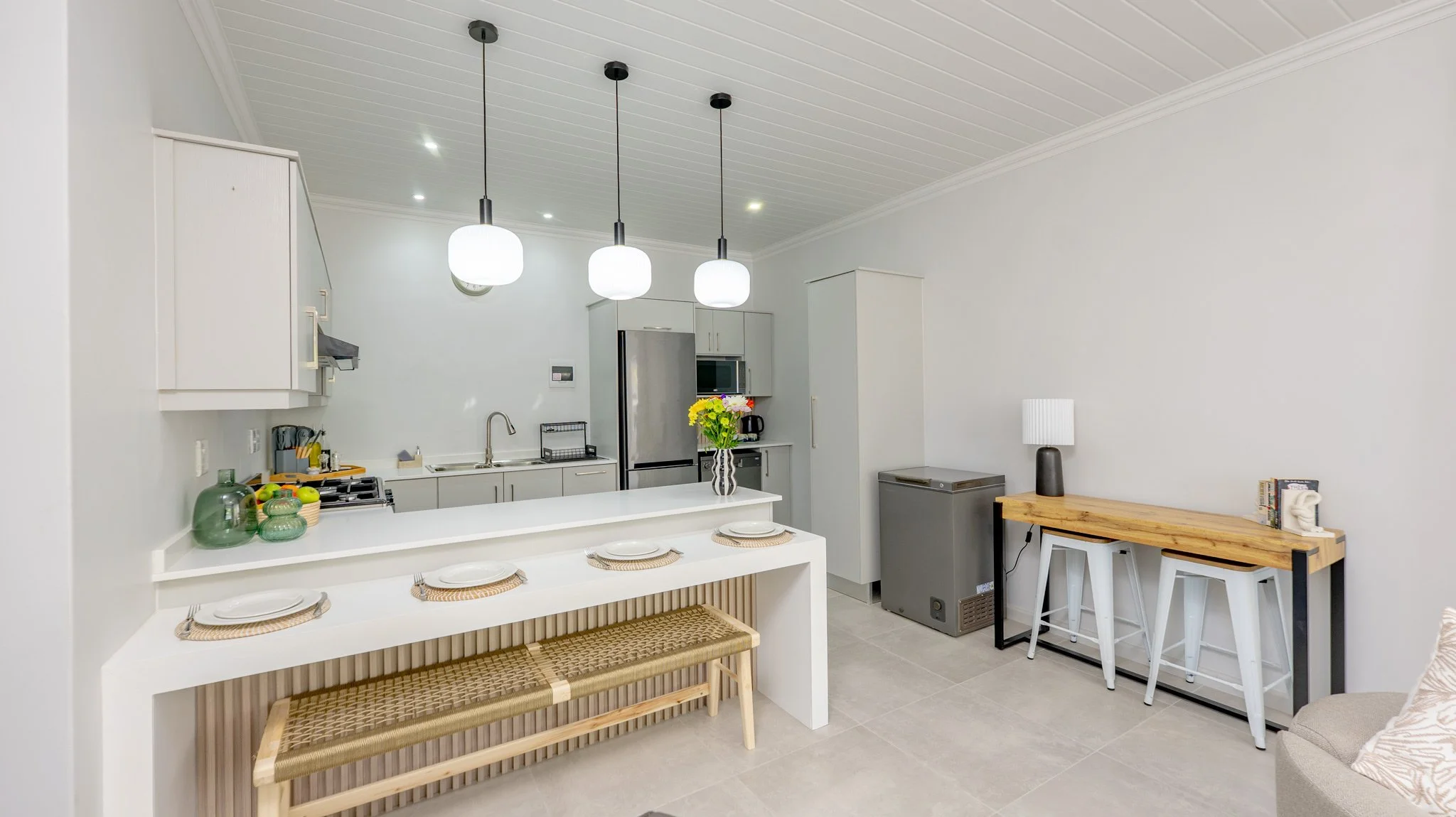 Modern open-concept kitchen and dining area with white walls, grey cabinetry, stainless steel appliances, and a white countertop island with four chairs. There are three pendant lights hanging over the island, a vase with flowers on the counter, and 