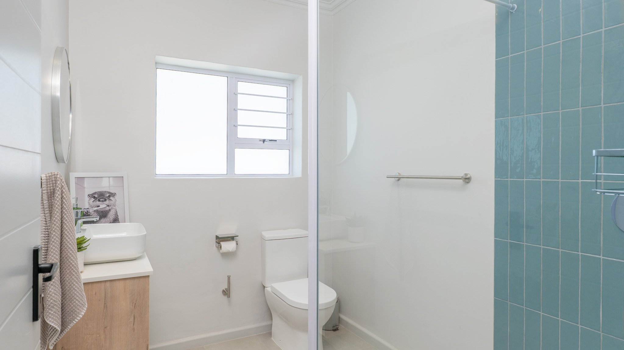A small bathroom with a white toilet, a window, a sink with a framed picture of a raccoon, a towel hanging on a hook, and a shower area with blue tiles.