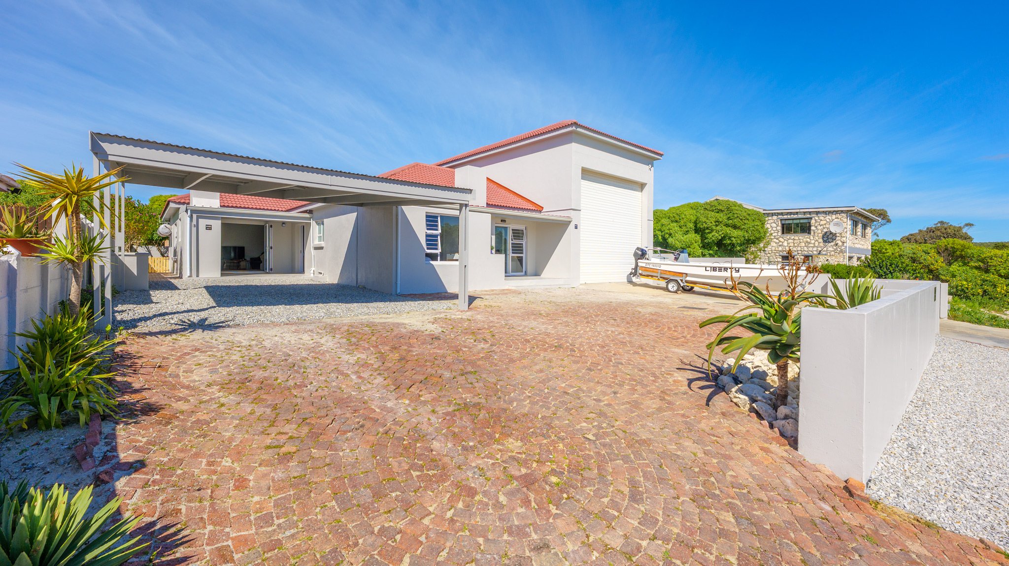 Modern house with white exterior, red roof, and a driveway with brick paving, surrounded by plants and greenery, under a clear blue sky.