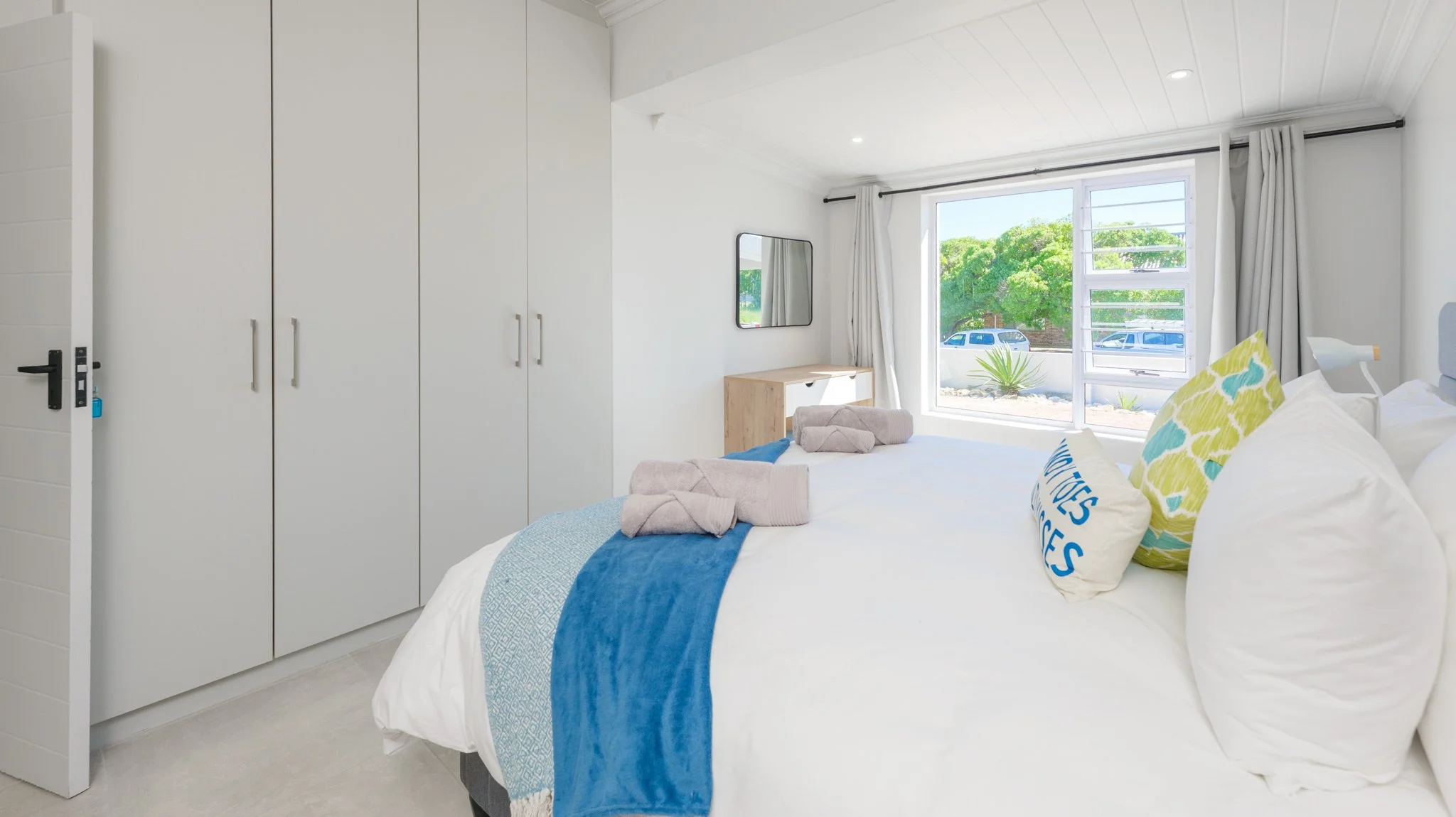 Bright bedroom with large window, white walls, and minimal decor. Bed with white linens, colorful pillows, and a blue throw blanket. Towels on the bed. Outside view of green trees and parked cars.