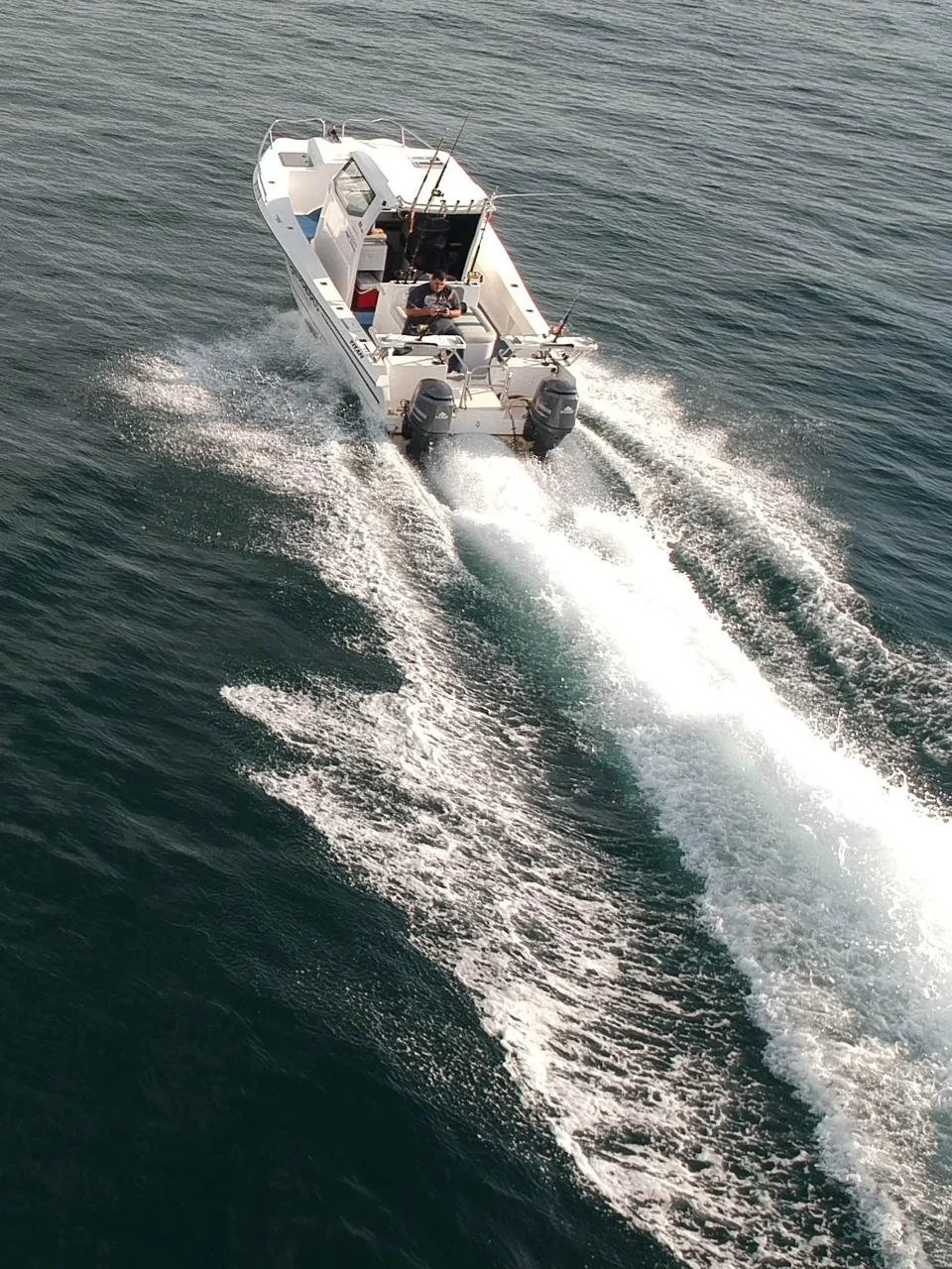 A white motorboat speeding on water, creating a wake behind it with two outboard engines, and a person fishing at the front.