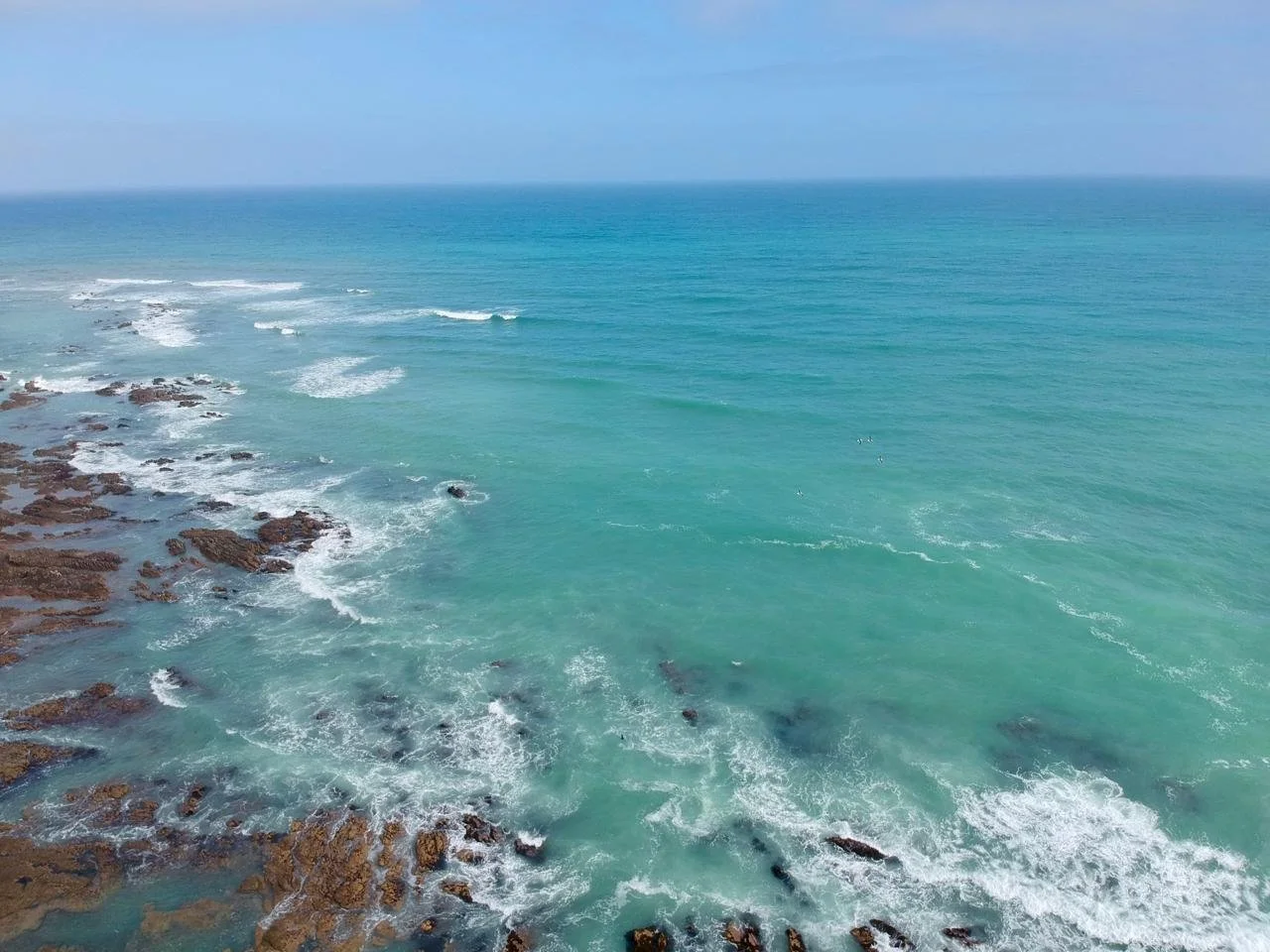 Aerial view of the ocean with turquoise water, white waves breaking on rocks, under a partly cloudy sky.