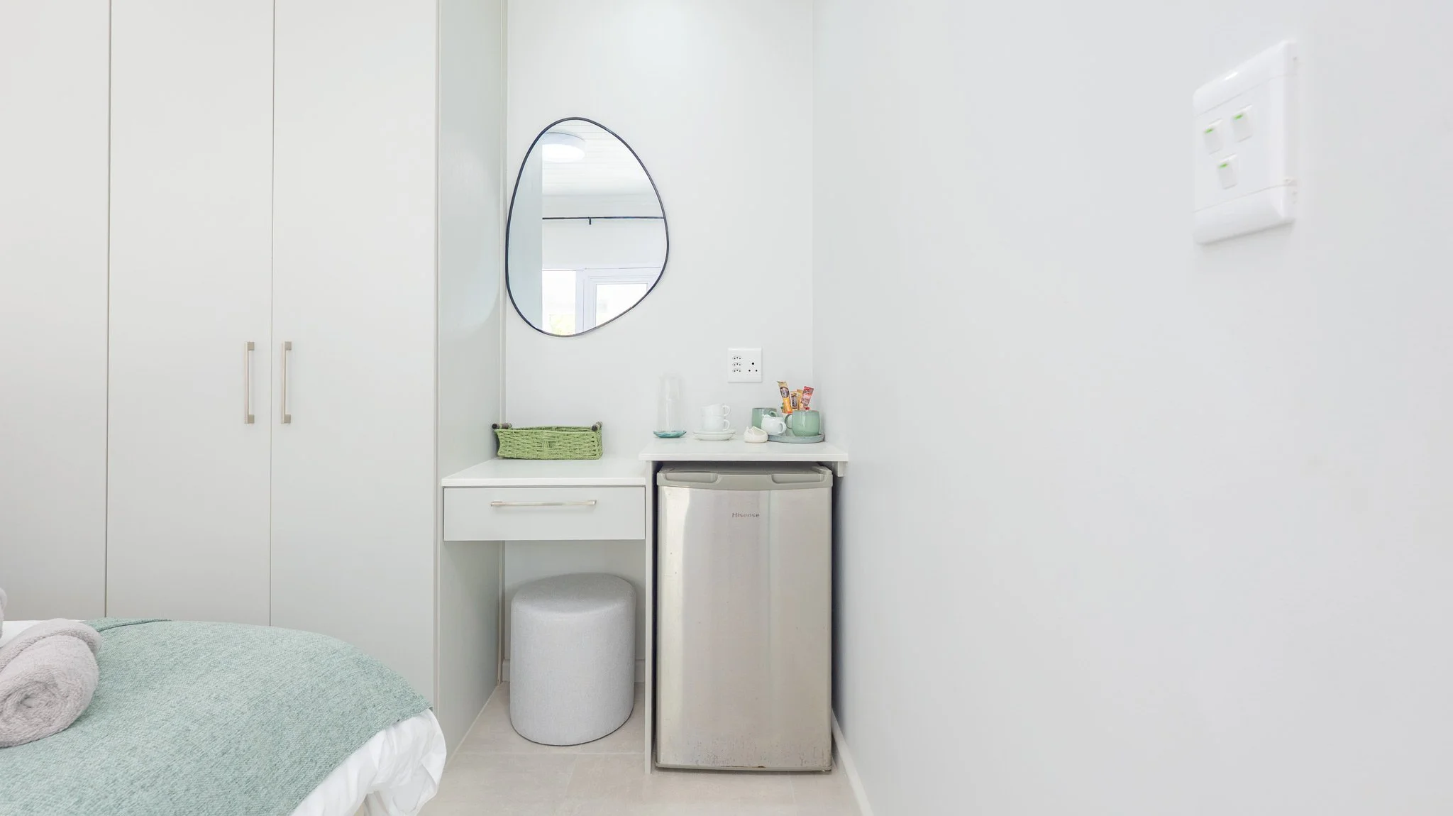 A small hotel room corner with a bed, white closet, mini fridge, and vanity with towel, mirror, and snacks.
