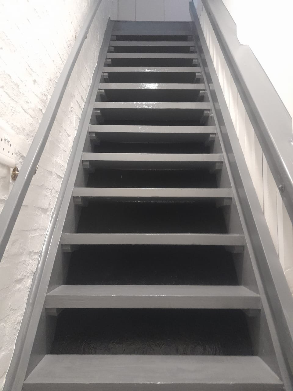 A metal staircase with black treads and grey risers, viewed from below, ascending against a white wall.
