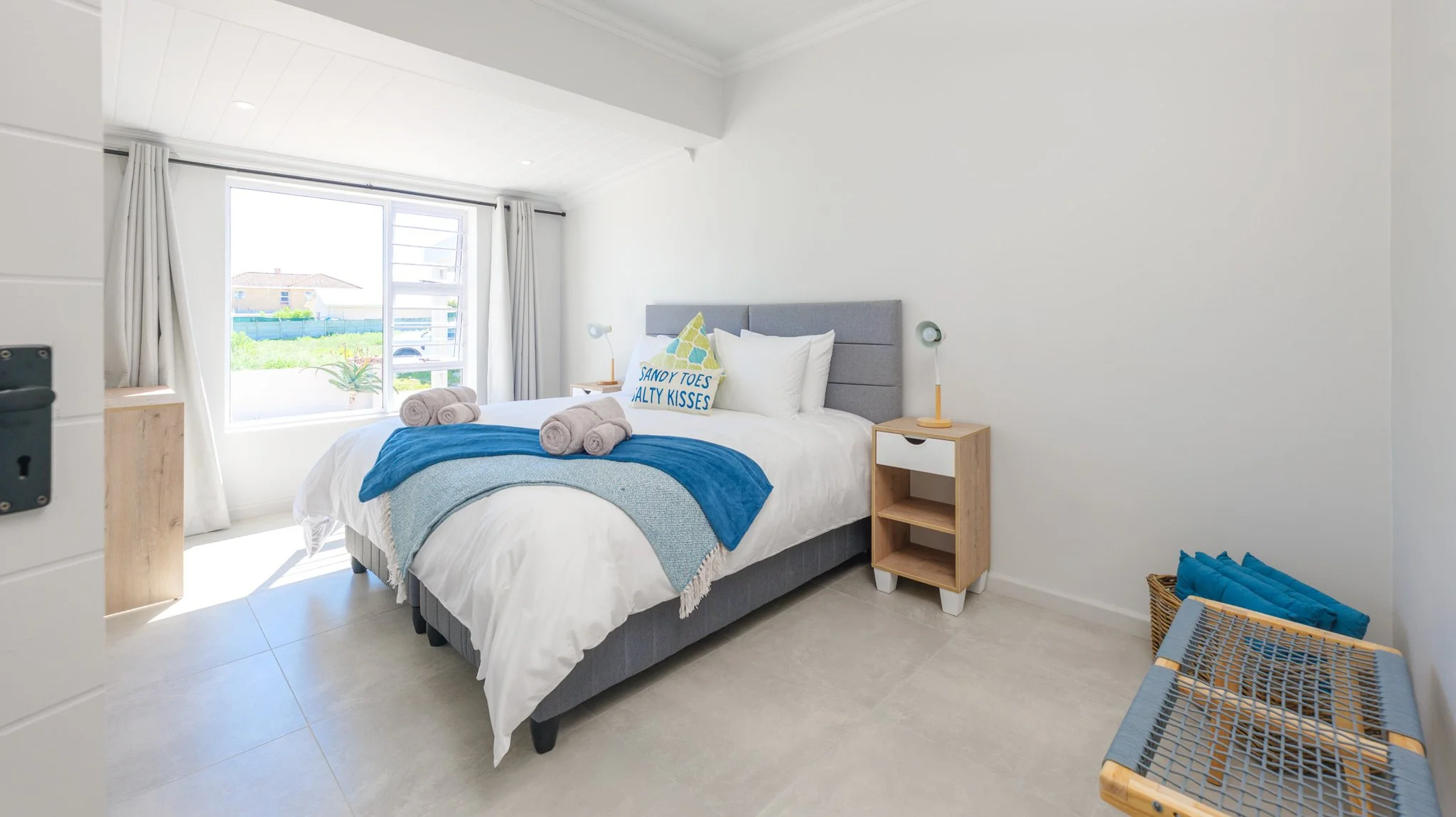 Bedroom with white walls, large window, bed with white bedding and blue accents, two rolled towels on the bed, decorative pillow, two bedside tables with lamps, wicker basket with blue pillows, and a small bench with a blue cushion.