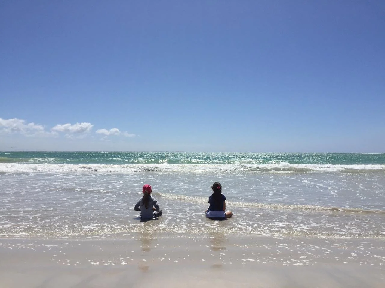 Two children sitting on the wet sand at the beach, facing the ocean, under a clear blue sky with minimal clouds.