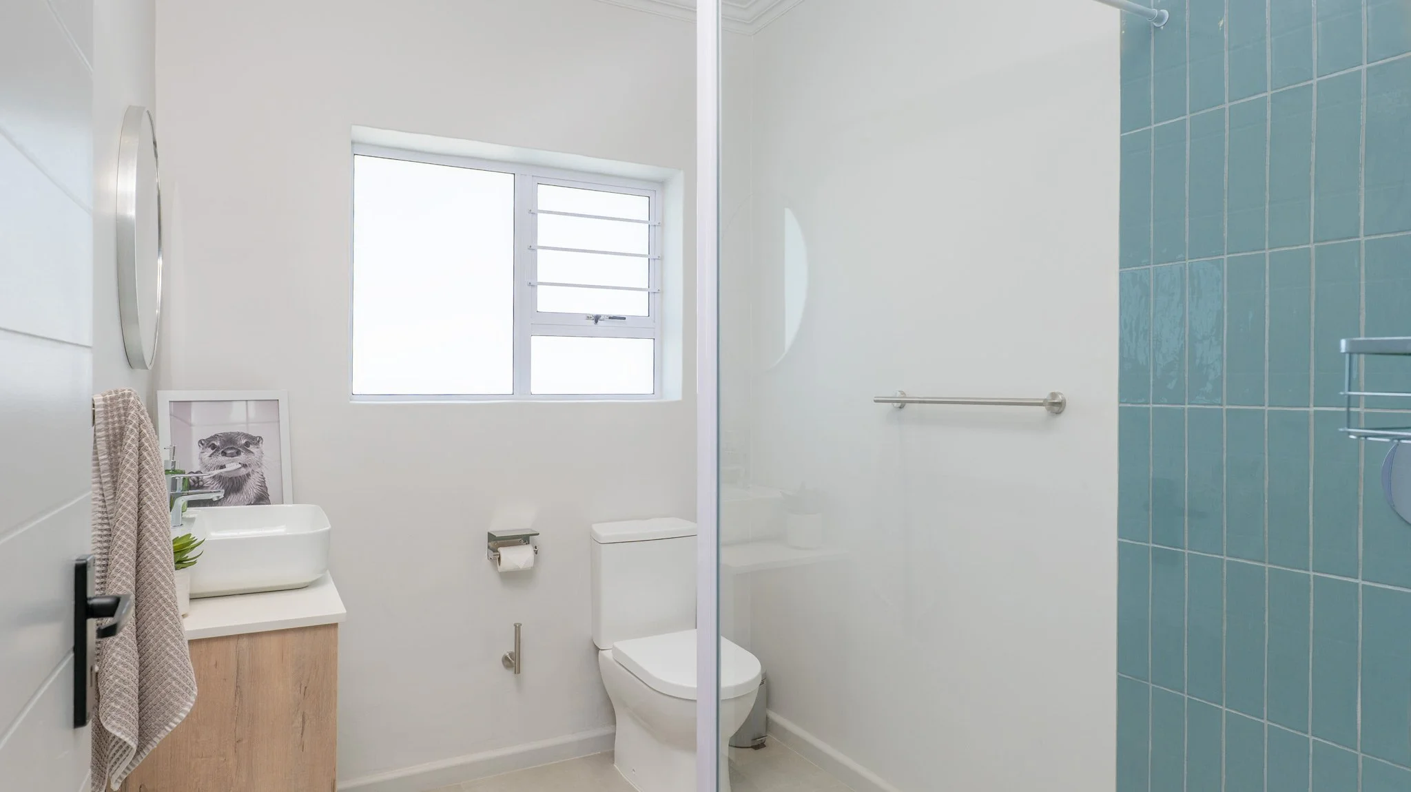 A small bathroom with a toilet, a window, and a shower area with blue tiles. There is a framed picture of a raccoon on the sink and a towel hanging on a hook.