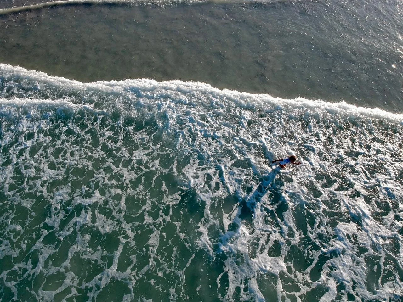 An aerial view of a person surfing in the ocean, with waves and foam surrounding them under sunlight.