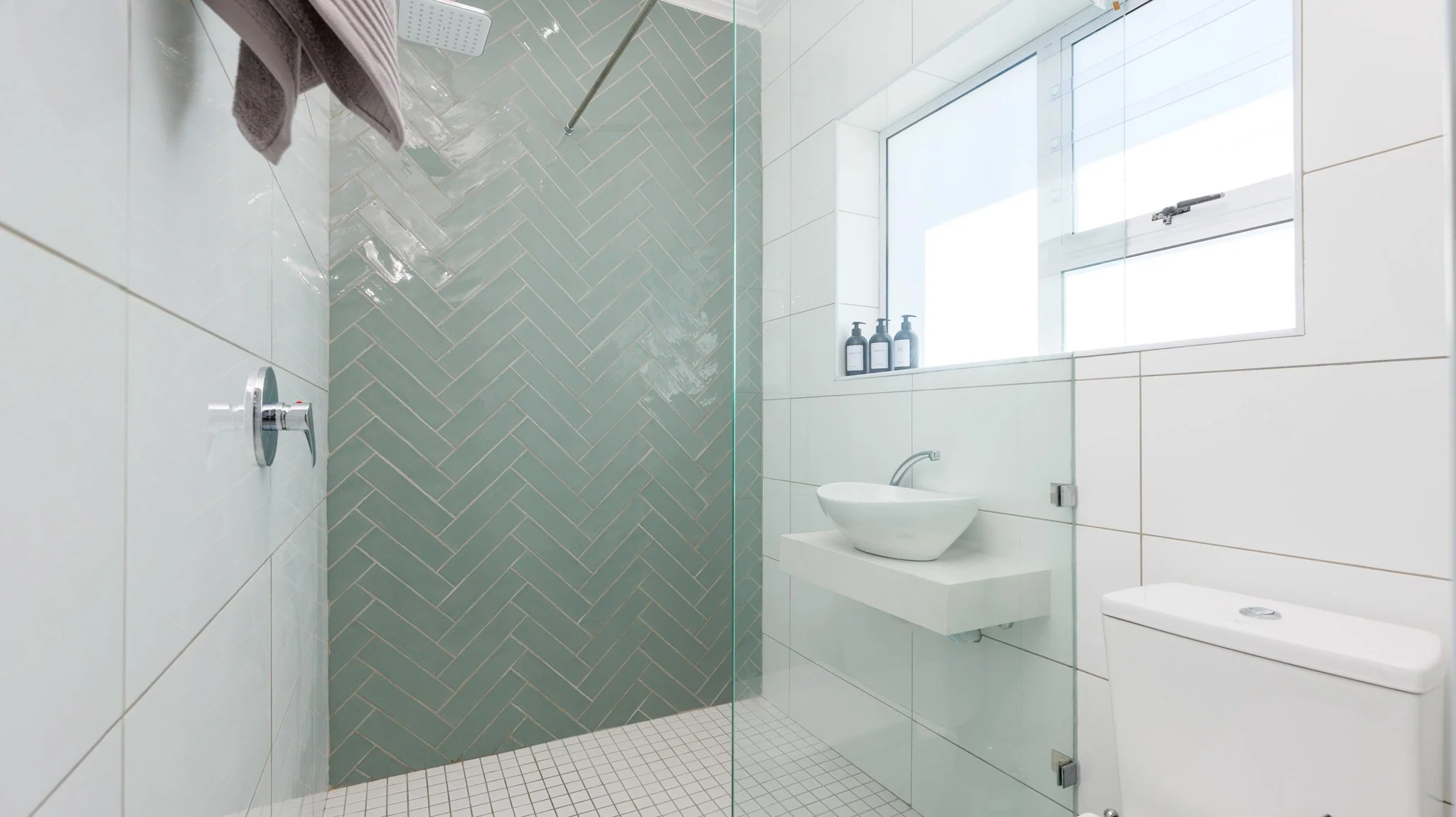 Modern bathroom with a walk-in shower featuring a green herringbone tile wall, a white sink on a floating counter, a toilet, and a large window with three bottles on the windowsill.