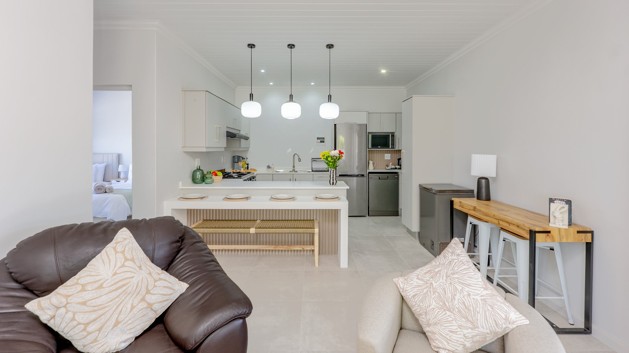 Open-plan living room and kitchen with gray walls, white cabinets, stainless steel refrigerator, microwave, and dishwasher, three pendant lights, a brown leather sofa with a cream pillow, a white armchair with a beige pillow, a wooden console table w