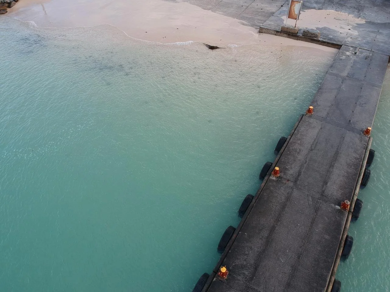 A concrete pier extending into turquoise water near a sandy beach with minimal waves. The pier has small orange safety lights and black tires attached along its sides.