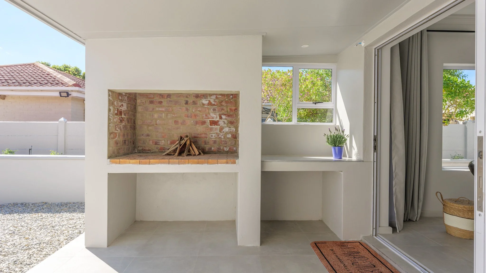 Outdoor patio area with built-in brick fireplace, white walls, window with green trees outside, potted plant, sliding glass door with curtains, and a doormat.
