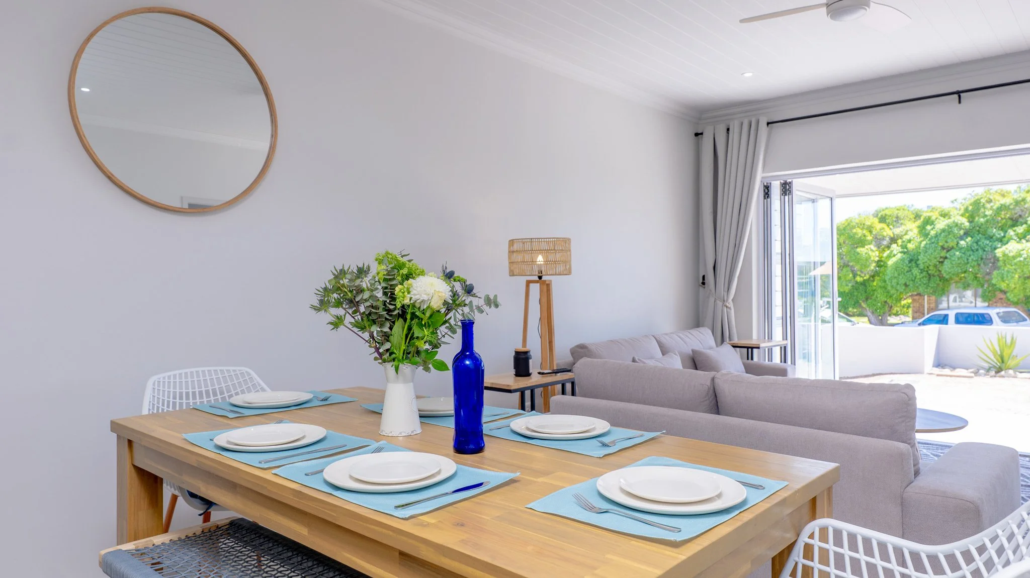 Modern dining and living room with a wooden table set with blue placemats, white plates, and a blue bottle with a flower arrangement. The living area has a beige sofa and a lantern-style table lamp near open glass doors leading outside, with greenery