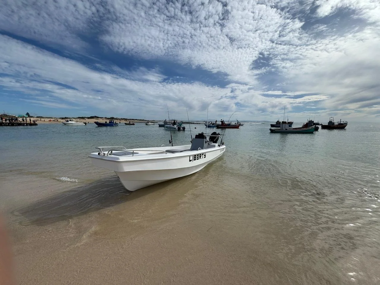 A white boat with the name LIBERTY on the side near the shore, with several other boats anchored in the water and a partly cloudy sky overhead.