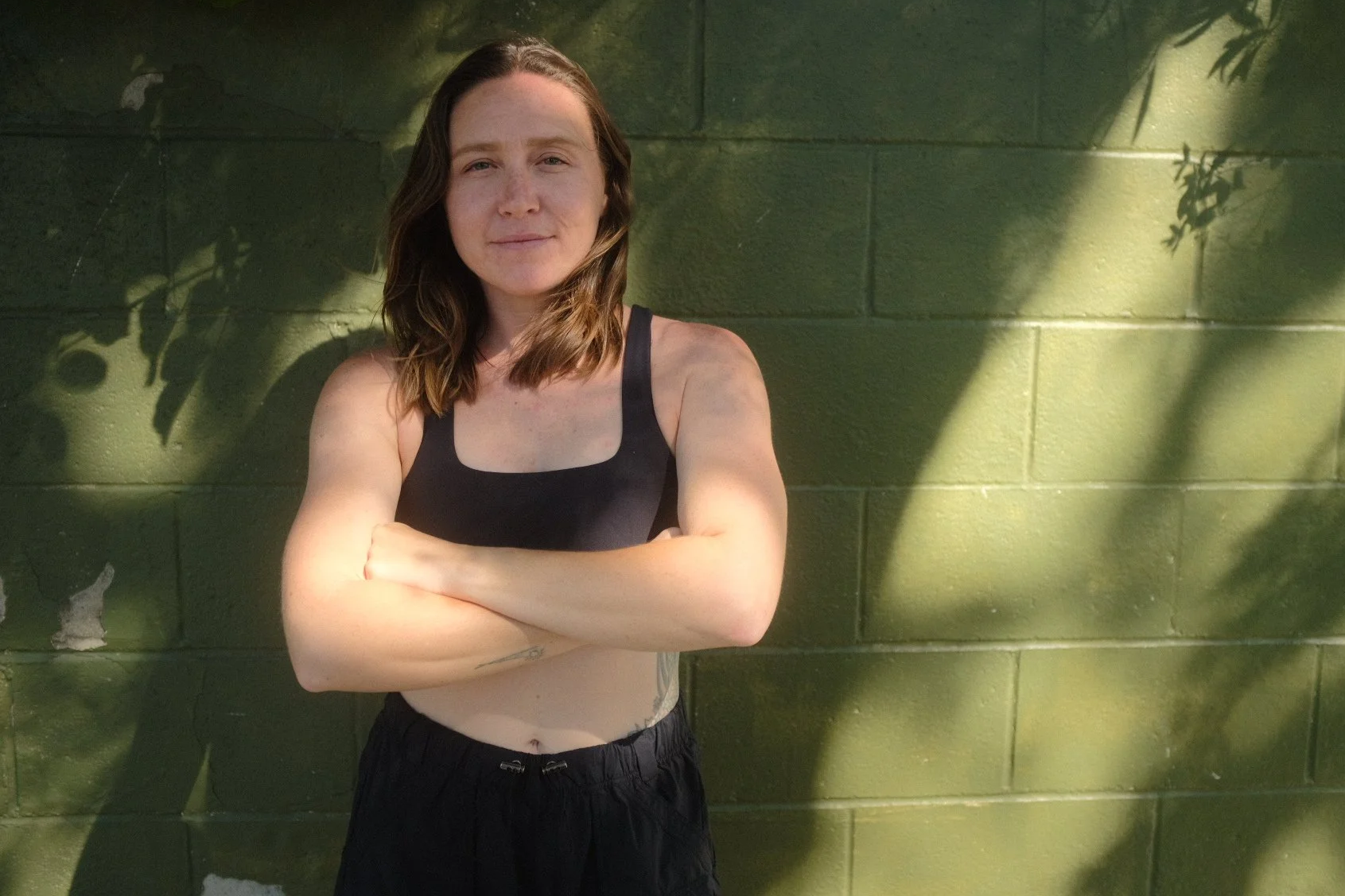 A woman with shoulder-length brown hair standing against a green painted brick wall, arms crossed, looking at the camera, with sunlight and shadows cast on the wall.