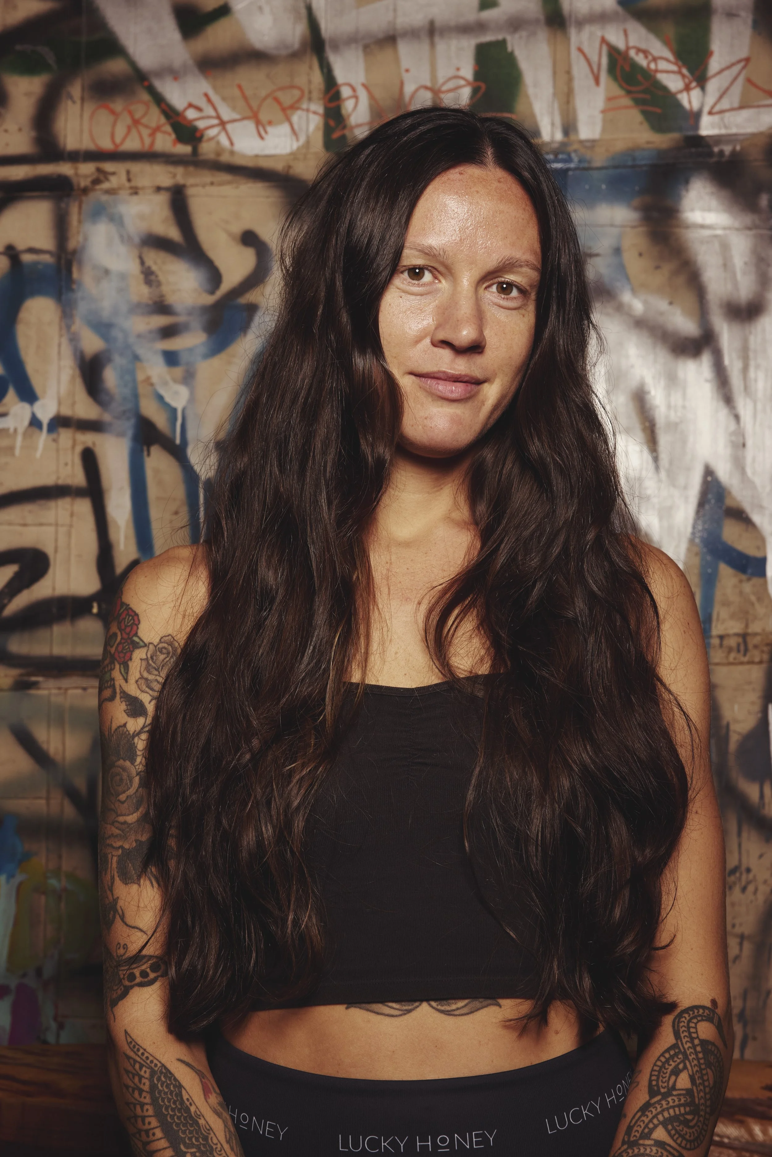 Woman with long braided hair, tattoos on arms and chest, wearing a black tank top, with hands pressed together in prayer, looking upward.