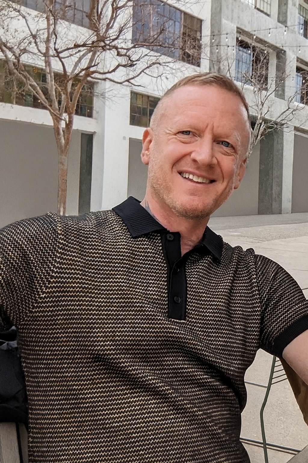 A man smiling outdoors in front of a modern building with large windows and leafless trees.