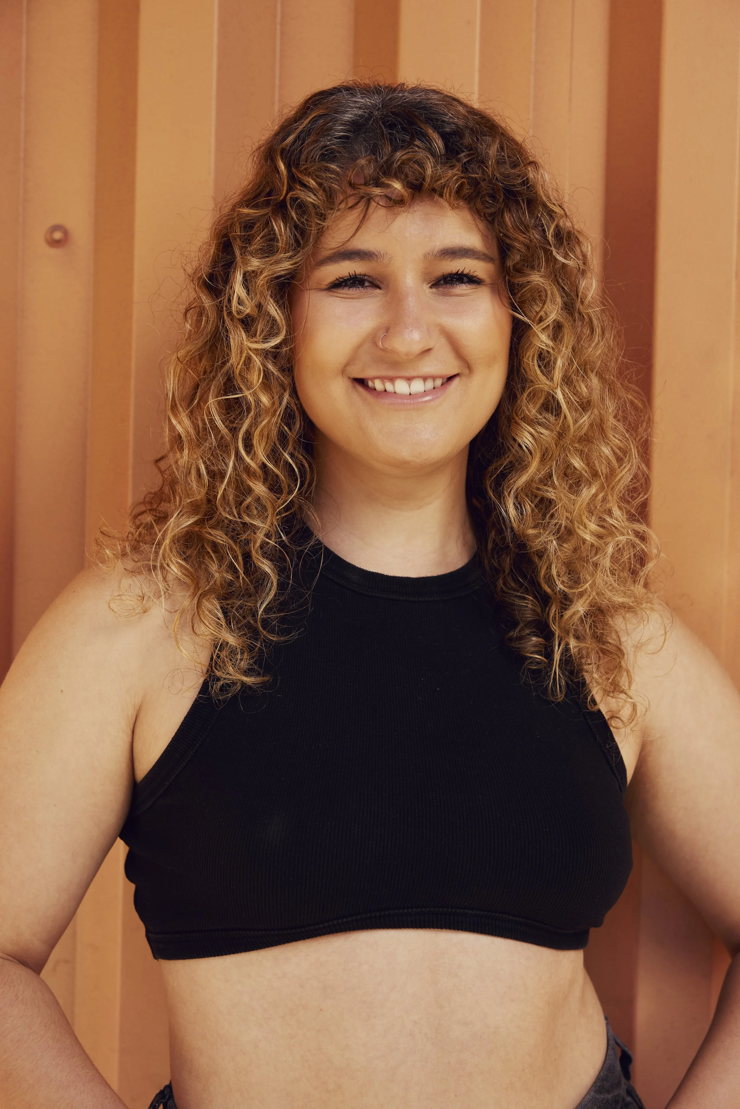 Portrait of a young woman with curly hair smiling, wearing a blue swimsuit, against a plain white background.