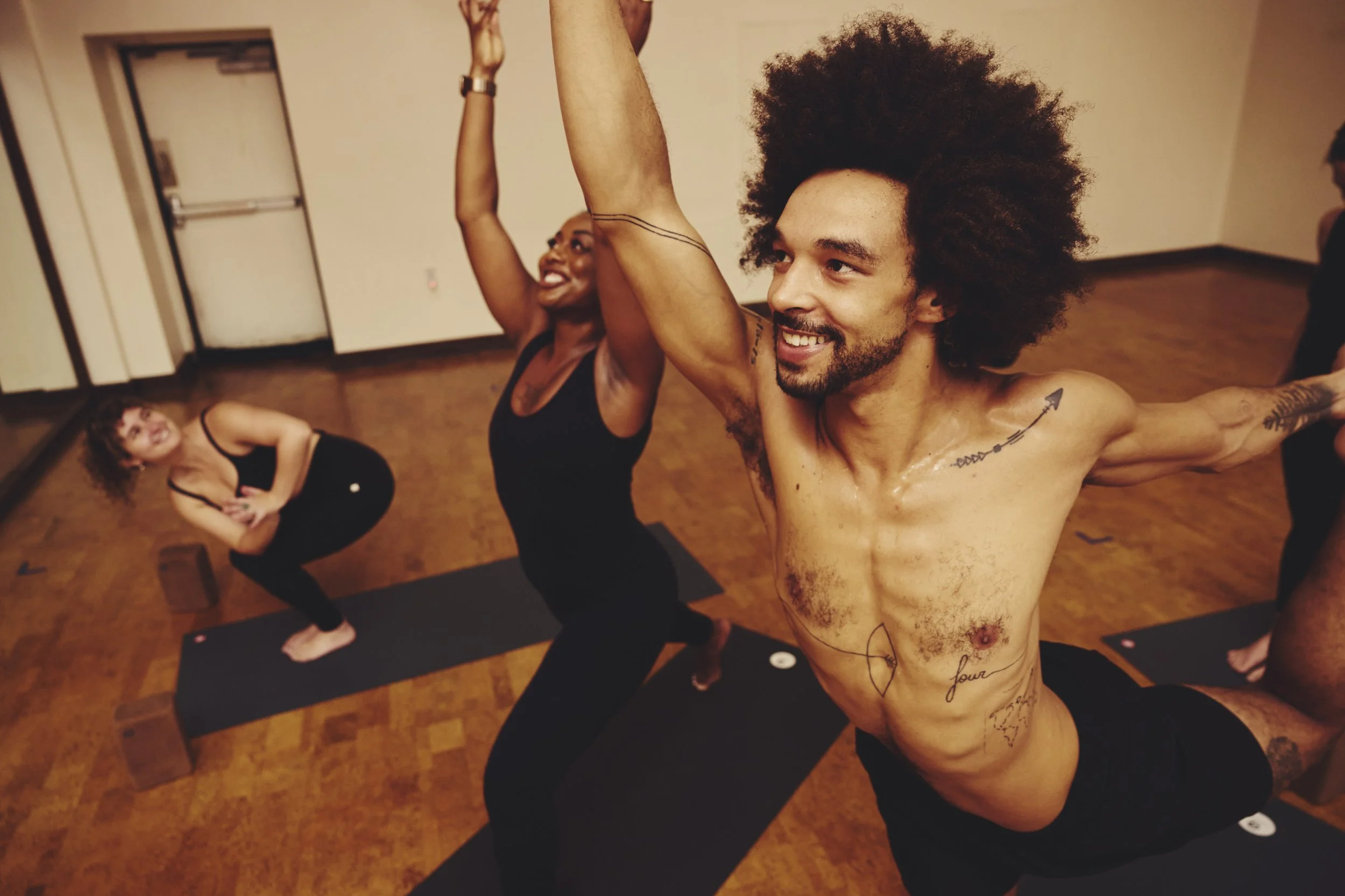 People practicing yoga in a studio with wooden floors. The man in the foreground has an afro and tattoos, smiling with arms raised. Two women in back are also performing yoga poses, smiling and looking happy.