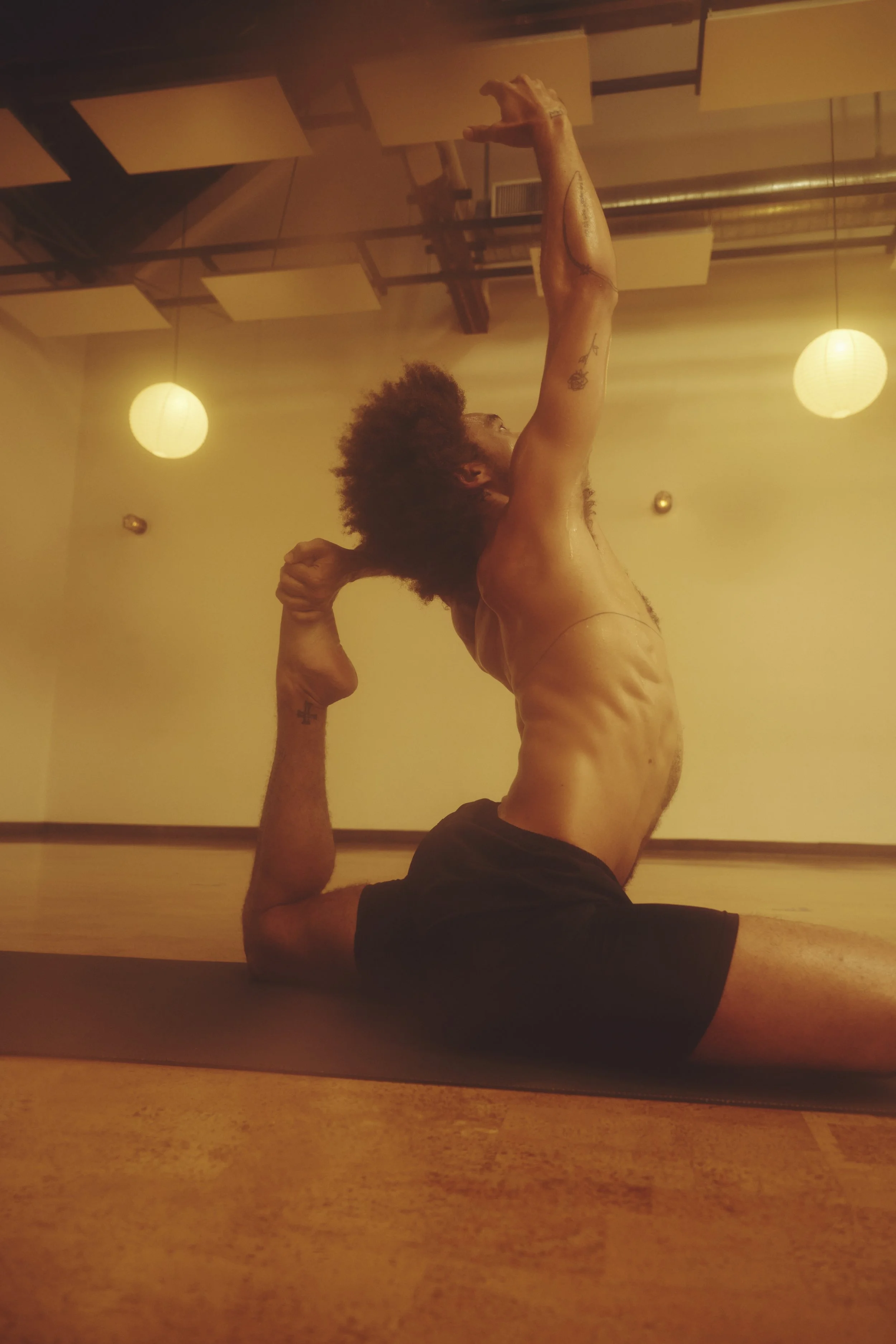 Person with curly hair practicing yoga in a studio, performing a pose with one arm extended upward and the other arm holding their foot behind them in a backbend, with warm lighting and hanging round lamps.