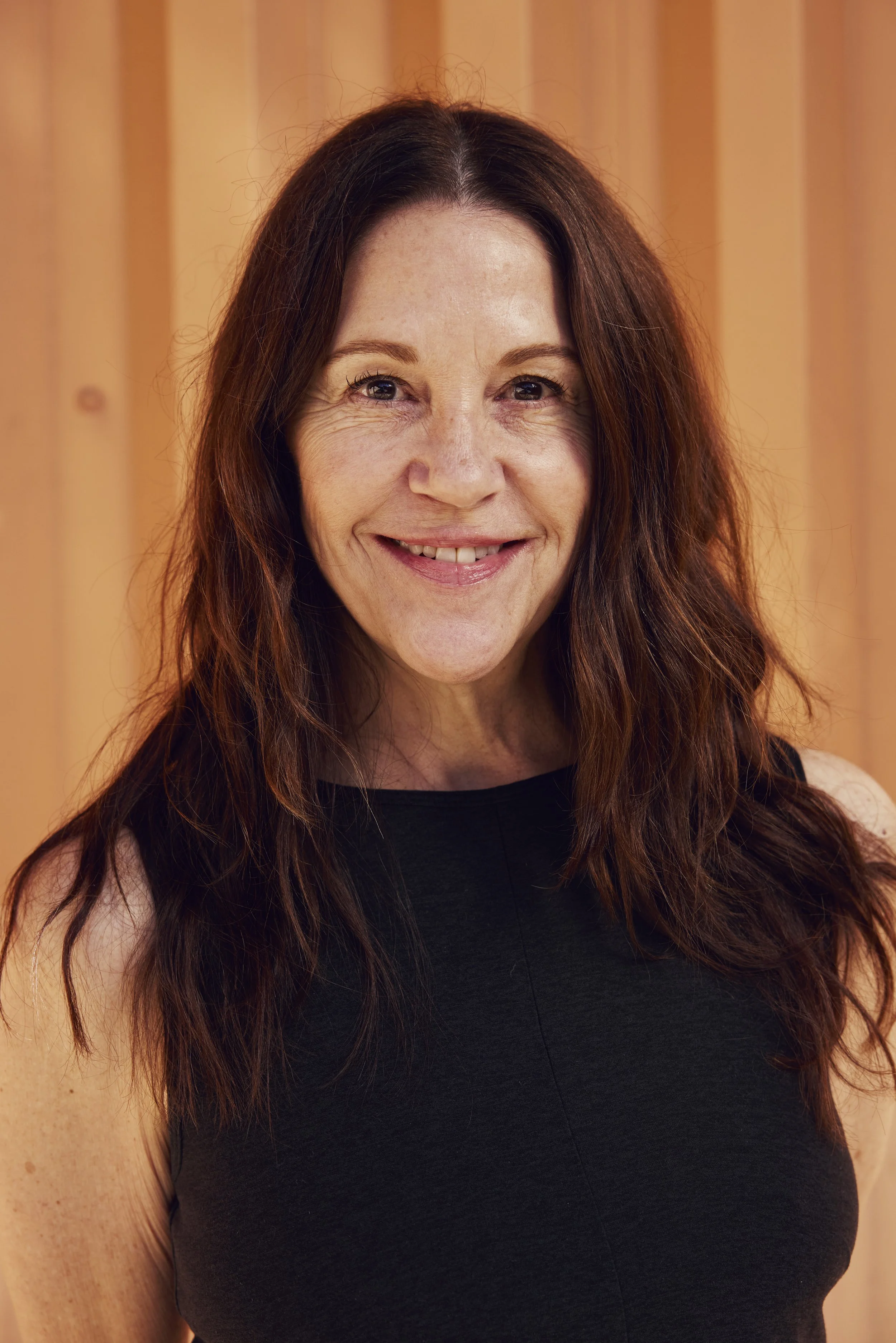 A woman with brown hair smiling and resting her arms on a table in an indoor setting with a blue poster or sign in the background and a green plant to the right.